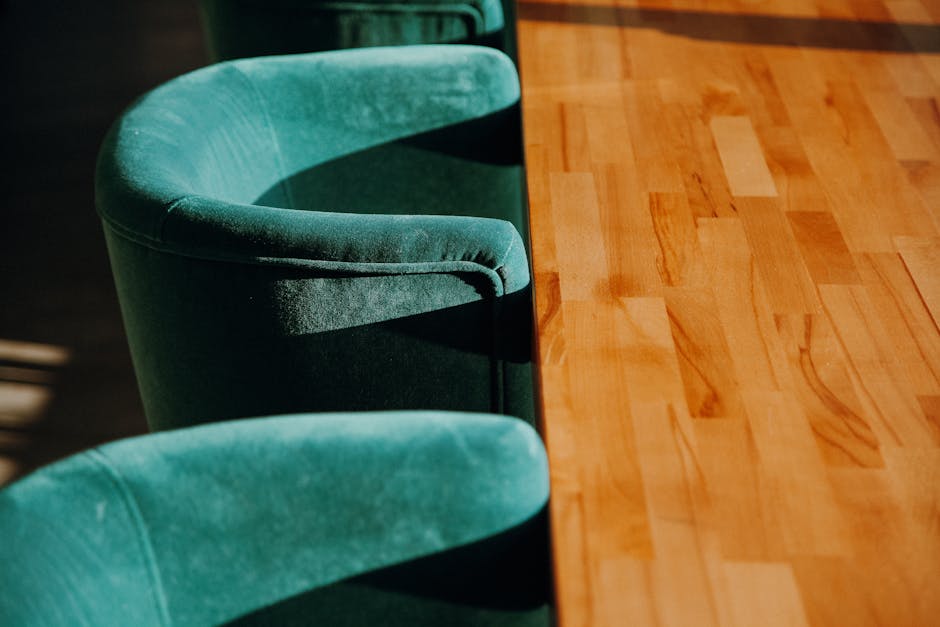 A close-up view of a polished wooden table with a warm, natural finish, positioned upright in a well-lit room. To the left, three green upholstered chairs with rounded backs and smooth fabric are arra